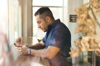 Man sitting at a long table in front of a window. He is looking at his phone.